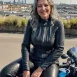 A smiling, middle-aged woman dressed in a black leather outfit, sitting on a motorbike.